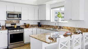 A modern kitchen with white cabinetry, stainless steel appliances, a granite countertop, and a dining area with white chairs. - Beautiful UPPER LEVEL Pet-Friendly Oceanfront Duplex P1111-1 (Oceanside)