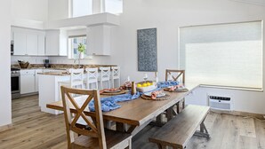 A modern kitchen with white cabinets, a dining area with wooden tables and chairs, and a large window with blinds. - Beautiful UPPER LEVEL Pet-Friendly Oceanfront Duplex P1111-1 (Oceanside)