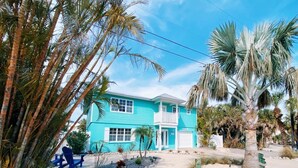 Een turquoise huis met twee verdiepingen en een balkon, omringd door palmbomen en een grindpad.