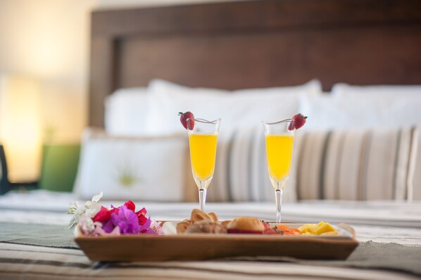 A breakfast tray with two glasses of orange juice, a plate of pastries, and a bowl of flowers on a bed with a white headboard.