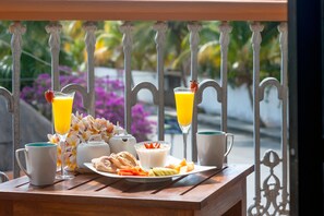 Une table de petit-déjeuner avec une assiette de nourriture, deux verres de jus d’orange, une tasse et une théière, le tout placé sur un balcon doté d’une balustrade en fer forgé et offrant une vue sur la verdure et les fleurs.
