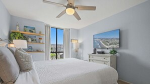 Une chambre à coucher avec un lit, un ventilateur de plafond, une télévision et une vue sur la ville par la fenêtre.