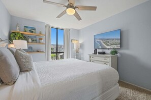 A bedroom with a bed, a ceiling fan, a TV, and a view of the city through the window. - Beach Theme Condo with Ocean & Pool View G222 North Coast Village (Oceanside)