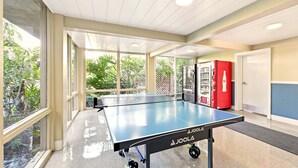 A ping pong table in a well-lit indoor space with large windows, a vending machine, and a door.