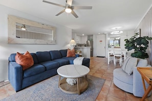 A living room with a blue sofa, a round coffee table, a ceiling fan, and a view into the kitchen.