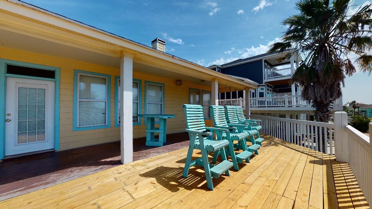 A wooden deck with turquoise chairs and a table, a yellow building with white trim, and a white railing.