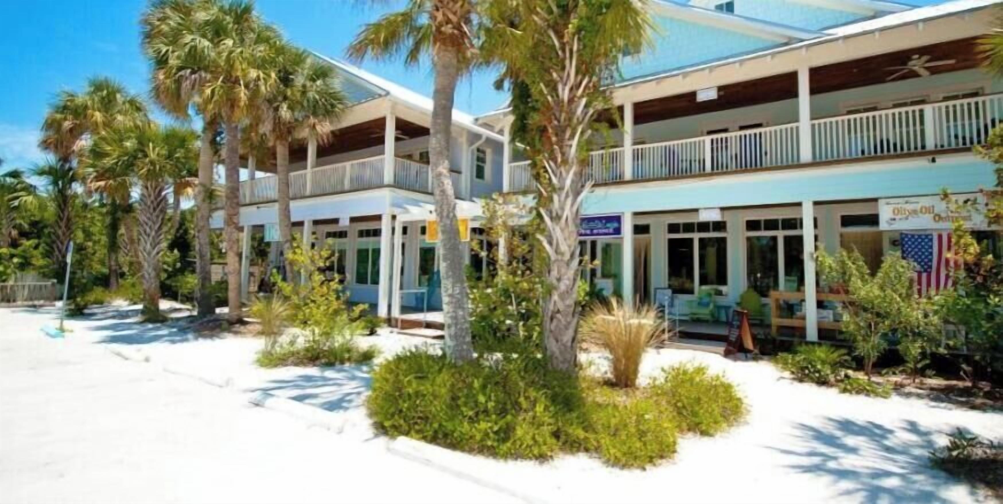 A two-story building with a balcony on the upper floor, surrounded by palm trees and a sandy area.