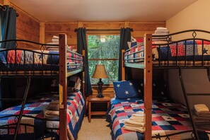 Uma cabana com três beliches, uma mesa pequena com um abajur e uma janela com cortinas.