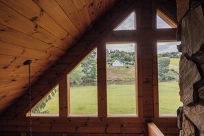 Quarto com teto inclinado e grandes janelas que oferecem vista para uma paisagem verde e um edifício branco ao longe.
