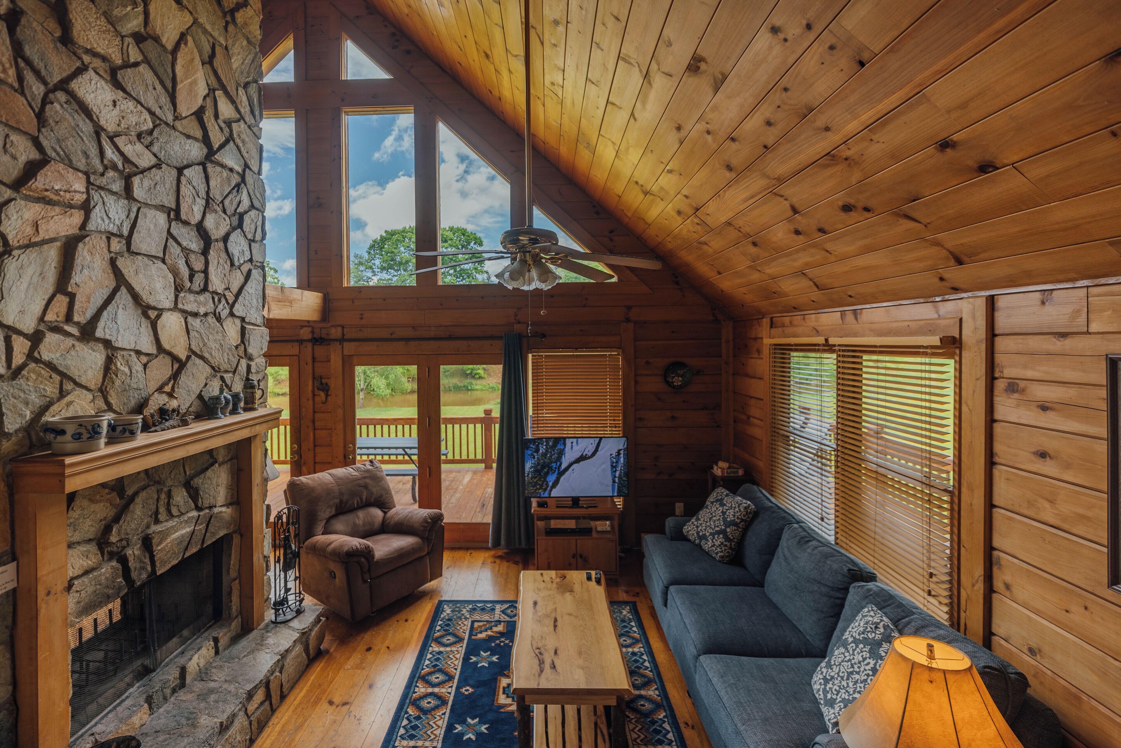 A cozy cabin interior with a stone fireplace, a wooden ceiling, and a large window overlooking a deck.