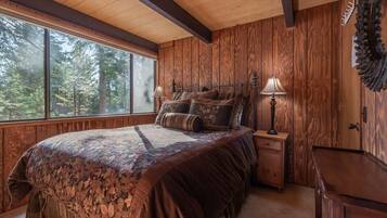 Ein Schlafzimmer mit einem großen Bett, Holzwänden, einem Fenster mit Blick auf einen Wald und einem Nachttisch mit einer Lampe.