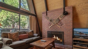 A cozy living room with a brick fireplace, a television, and a large window overlooking a forest. - Unique 3 Story with Two Great Entertainment Areas! Short drive to Lake and Northstar! (Kings Beach)