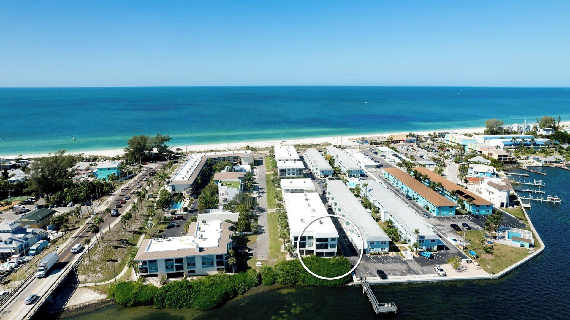An aerial view of a coastal area with a marina, a beach, and a row of buildings with parking lots.