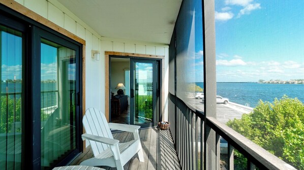 A balcony with a white chair, a glass door leading inside, and a view of the sea and a parking area.