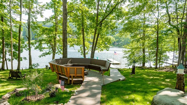 A lakeside deck with a curved bench and a small American flag, surrounded by lush greenery and a view of the lake. - Dog Friendly Getaway Just Steps to Wisp & Fun (MCHENRY)