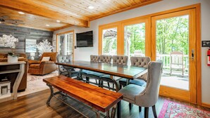 A spacious room with a large wooden dining table, comfortable seating, a television, and large windows offering a view of the outdoors. - Dog Friendly Getaway Just Steps to Wisp & Fun (MCHENRY)