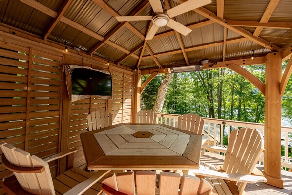 A wooden deck with a table and chairs, a ceiling fan, and a view of a lake surrounded by trees.
