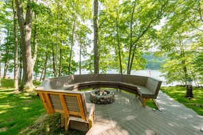 A wooden deck with a built-in fire pit and seating area surrounded by trees, overlooking a body of water.