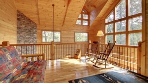 A wooden cabin interior with a stone fireplace, a patterned sofa, a rocking chair, and large windows offering a view of the forest. - CHEROKEE DREAM - SECLUDED MTN VIEW, POOL TABLE, AIR HOCKEY, FIRE PIT, SCREENED PORCH (Epworth)