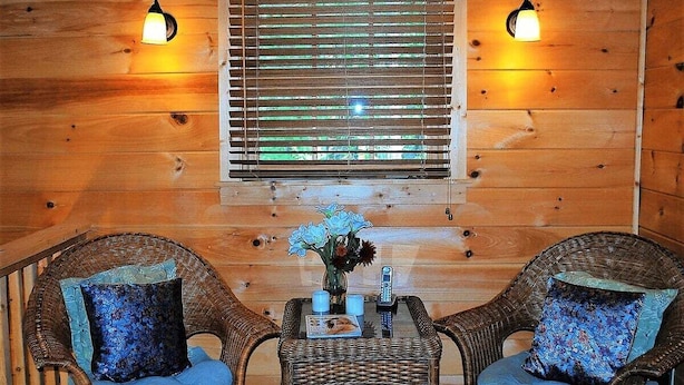 A cozy room with wooden walls, two wicker chairs with blue cushions, a small table with a vase of flowers, and two wall-mounted lamps.