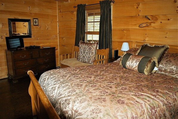A wooden cabin room with a bed, a dresser, a chair, and a window with curtains.