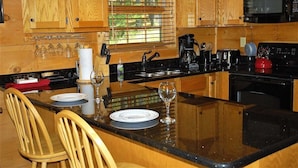 A kitchen with wooden cabinets, a black countertop, a sink, a stove, and a microwave. There are two chairs and a table with wine glasses and plates.