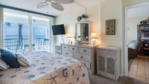 Ein Schlafzimmer mit einem großen Bett, einem Deckenventilator, einem Fernseher, einer Kommode und Blick auf das Meer durch die Schiebeglastüren.