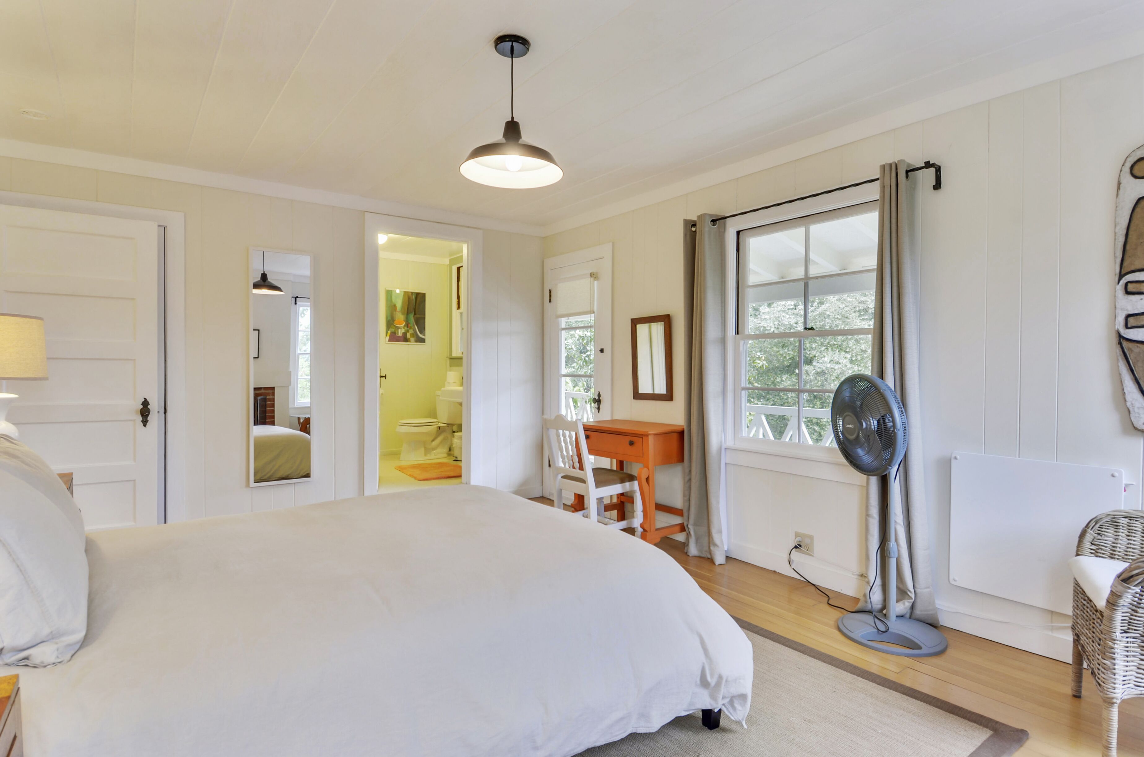 A bedroom with a large bed, a desk, a chair, a standing fan, and a window with curtains.