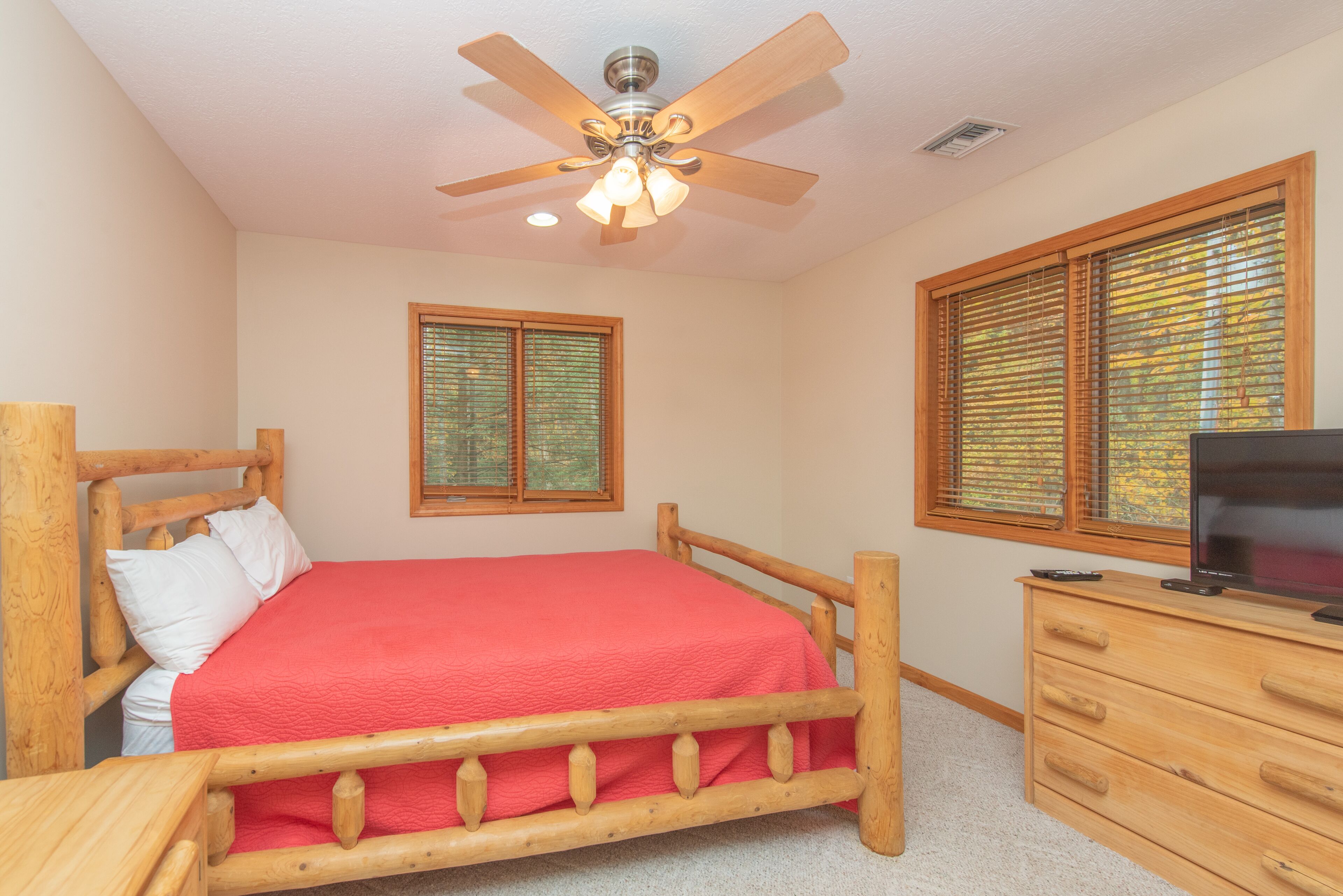 A bedroom with a wooden bed, a ceiling fan, a TV on a wooden dresser, and two windows with blinds.