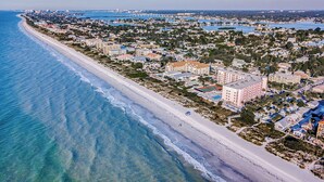 An aerial view of a coastal town with a long sandy beach, residential buildings, and a large hotel with a prominent building in the center. - Gulf Front BEACH getaway - Reef Club 308 (Indian Rocks Beach)