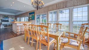 A spacious dining area with a glass-top table and wooden chairs, large windows offering a view of the beach, and a chandelier above. - Timeless Oceanfront Beach Retreat in DeBordieu (Georgetown)
