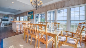 A spacious dining area with a glass-top table and wooden chairs, large windows offering a view of the beach, and a chandelier above.