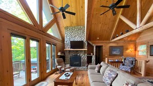 A cozy cabin interior with a stone fireplace, wooden ceiling beams, and a large window overlooking a deck. - Mountain Cabin,Pool Table,Views,Hot Tub,Fireplace, WiFi, Pet Friendly (Ferguson)