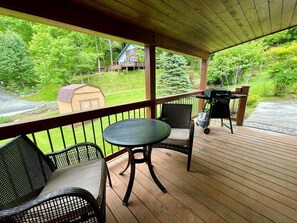 A covered outdoor area with a round table and two chairs, a grill, and a view of a house and greenery. - River Hill - River Access, Dog Friendly, Pool Table, WIFI, River View, Gas Fireplace, Mtn Views (Crumpler)