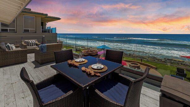 Een balkon met rieten meubels, een glazen tafel en uitzicht op de oceaan bij zonsondergang.