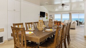 Een eethoek met een houten tafel, stoelen en gevlochten manden. Grote ramen bieden uitzicht op de oceaan.