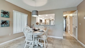 A dining area with a round table and white chairs, a pendant light, and a view into the kitchen. - Gulf Watch 111 - Anna Maria Island - Condo 2 Bedroom / 2 Bath Gulf to Bay access, maximum occupancy of 5 people. (Bradenton Beach)