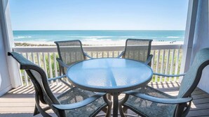 Un espace extérieur abrité, doté d’une table en verre et de chaises, offrant une vue dégagée sur une plage et l’océan.