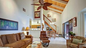 A spacious living room with a high ceiling, a large ceiling fan, and a view of the kitchen through an open door.