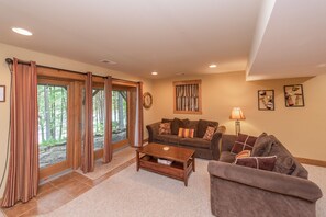 A living room with a brown sofa, a wooden coffee table, and a lamp. There are large windows with curtains and a view of trees. - Cozy Lakefront Charm w/ Dock & Hot Tub (SWANTON)