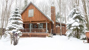 Une cabane en bois dotée d’un porche et d’une cheminée, entourée d’arbres couverts de neige.