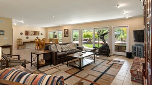 A spacious living room with a sofa, coffee table, and chairs, a dining area with a table and chairs, a treadmill, and a TV.