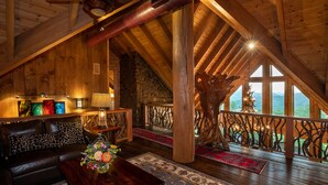Un acogedor dormitorio tipo loft con techo de madera, una chimenea de piedra, una escalera de madera y un balcón con vistas a las montañas.