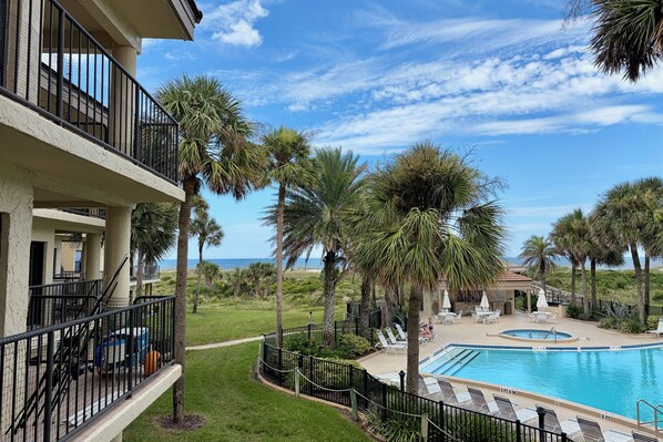 A pool area with a view of the ocean, surrounded by palm trees and a building with balconies.