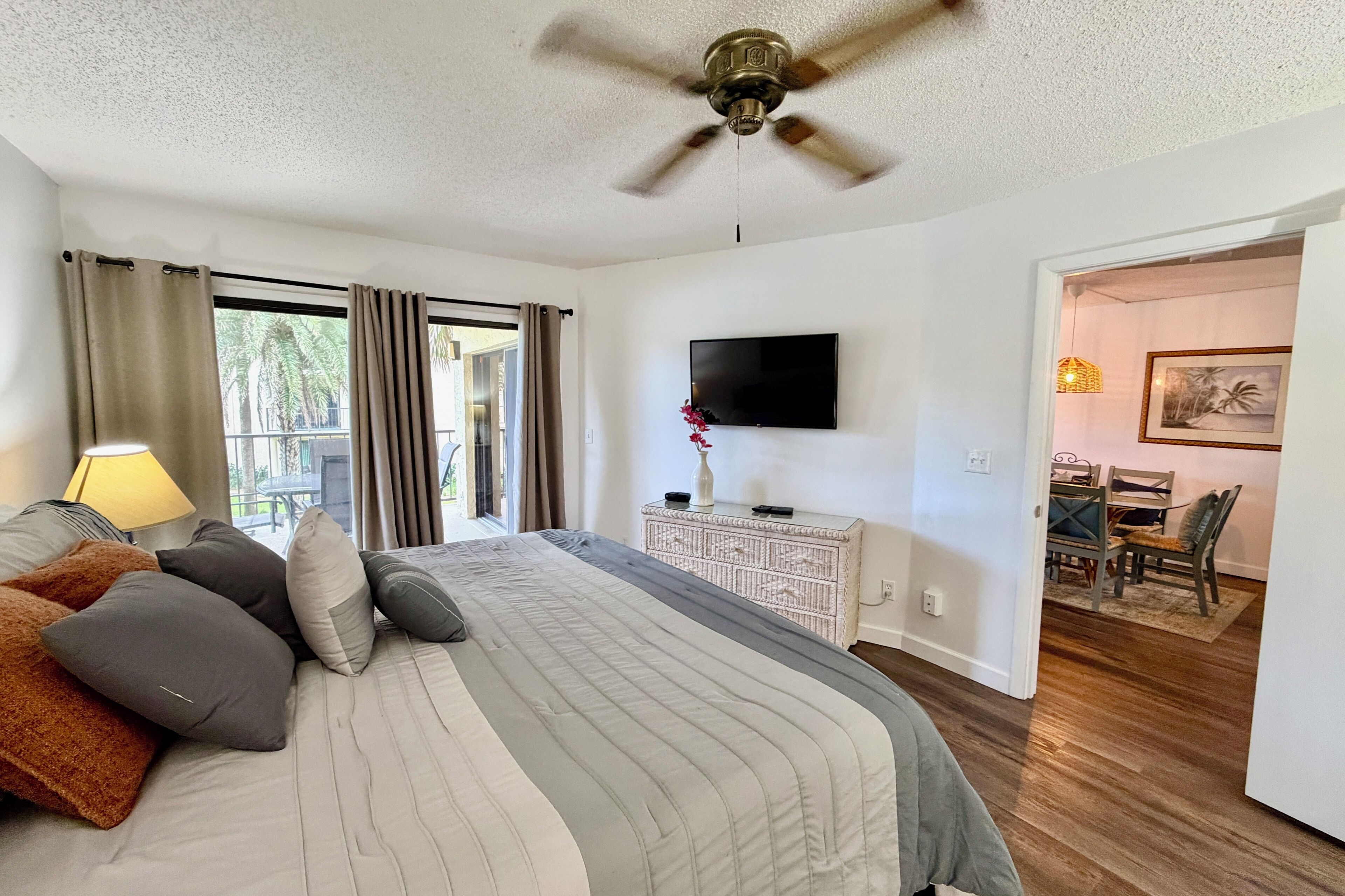 A bedroom with a bed, a ceiling fan, a TV, a dining area with chairs, and a view of palm trees outside.