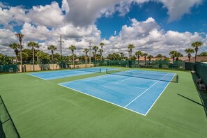 Un court de tennis extérieur doté de plusieurs terrains, entouré de palmiers et surplombé d’un ciel dégagé.