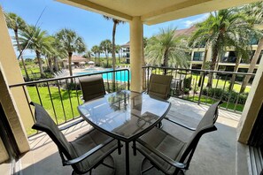 Un balcón con una mesa y sillas de vidrio, con vista a una piscina y palmeras.