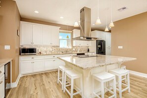 A kitchen with white cabinets, a central island with stools, stainless steel appliances, and pendant lighting. - The Flight Deck: 4 BR, 3 BA House in Port Bolivar, Sleeps 12 (Bolivar Peninsula)