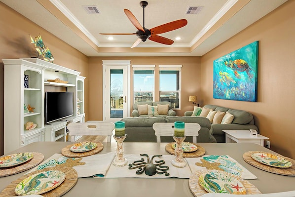 A living room with a ceiling fan, a television, a sofa, and a dining table set with plates and candles. - The Flight Deck: 4 BR, 3 BA House in Port Bolivar, Sleeps 12 (Bolivar Peninsula)