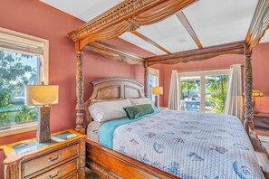 A four-poster bed with a floral bedspread, a wooden nightstand with a lamp, and a view of the outdoors through a sliding glass door.
